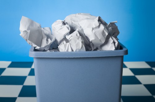 A close-up view of a light blue plastic rubbish bin filled with crumpled sheets of white paper, positioned on a black and white checkered floor. The background features a plain blue wall, suggesting an indoor environment possibly within an office or storage area. The crumpled paper appears used and discarded, with some sheets sticking out from the top of the bin. The texture of the paper is visibly wrinkled, and the bin's smooth surface contrasts with the paper's crumpled appearance. The overall scene reflects a typical waste collection or rubbish removal context, aligned with services offered by Office Clearance Mottingham on the postcode area relevant to the town, with the bin placed centrally to emphasize the disposal of paper waste commonly encountered during office clearances or rubbish disposal tasks in the local area.