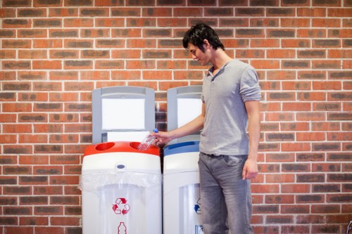 A young man with dark hair, dressed in a light grey long-sleeved shirt and grey jeans, is standing outdoors against a red brick wall, holding a plastic bottle in his right hand, preparing to discard it in a white recycling bin with a red recycling symbol on the front. Adjacent to him is a second white bin with a blue recycling symbol, indicating separation of waste types. The bins are positioned on a paved surface, and the scene appears to be part of an urban or semi-urban setting, possibly near an office or residential area in the UK. The image visually relates to rubbish disposal and recycling services offered by companies like Office Clearance Mottingham, emphasizing environmentally responsible waste management solutions in the local area.