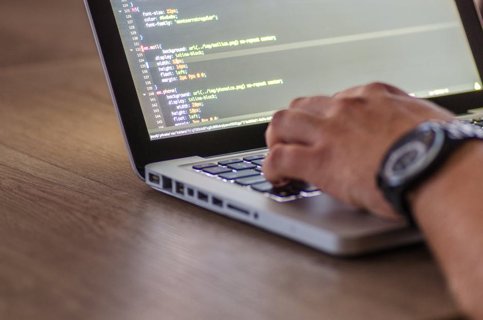A person with short dark hair, seen from behind, is seated at a desk working on a laptop computer. The laptop's screen displays lines of colorful code on a dark background, indicating programming acti