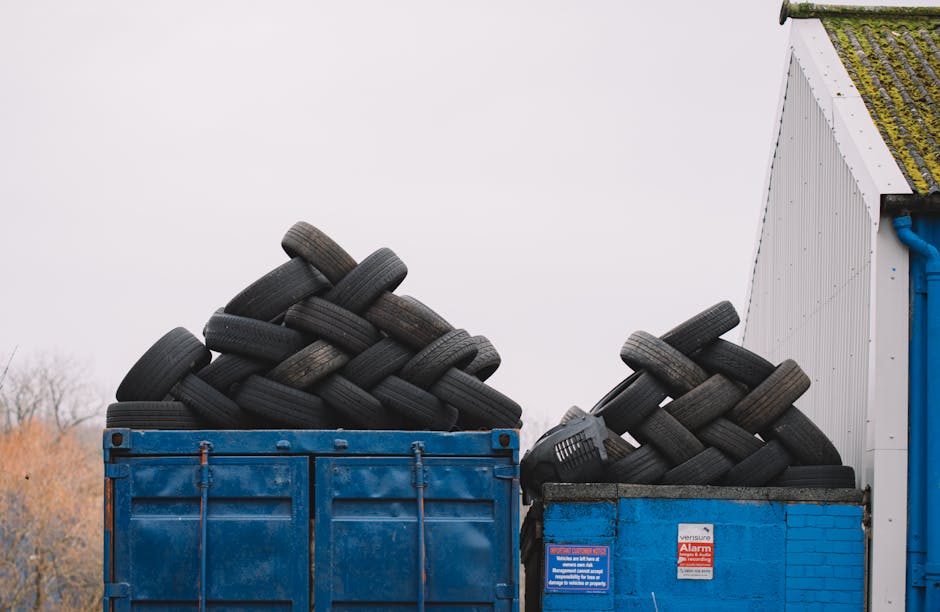 A large blue metal container filled with stacked black rubber tires sits outdoors against a cloudy sky. The tires are piled unevenly, with some leaning on each other and others stacked horizontally, d