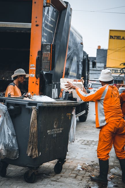 A large pile of mixed rubbish including cardboard boxes, plastic bags, paper waste, and various discarded items, located on a paved pedestrian area in front of a row of recycling bins. The rubbish ext