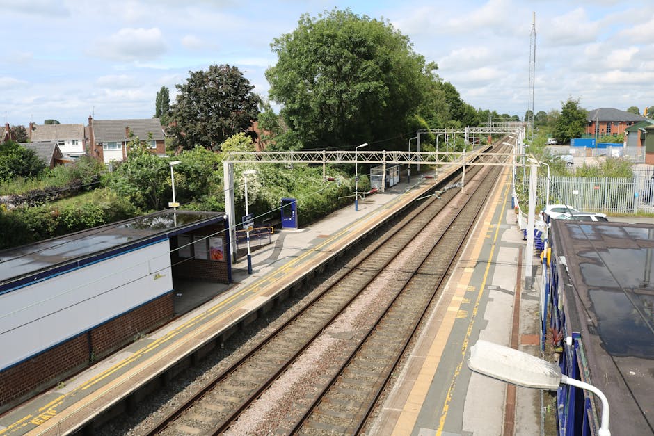 An elevated view of a railway station platform with two train tracks running parallel, flanked by yellow safety lines and bordered by a small brick shelter with a black roof on the left side. The shel